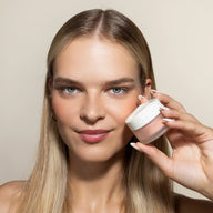A blonde woman holding a sUoga Uoga 643 Peach Blush Powder jar next to her cheek, showing a close-up of her face and the product against a plain beige background.