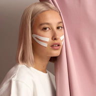 Woman wearing Lyfta white face lifting kinesiology tape with white face paint against pink curtain
