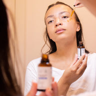 Woman getting an eye contour serum applied under her eye with a dropper by another person, holding a Clémence & Vivien bottle in the foreground while a second bottle is visible in the background.