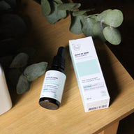 Clémence & Vivien Huile de Soin Peaux Mixtes bottle and box lying on a wooden shelf beside eucalyptus branches, moody dark background.