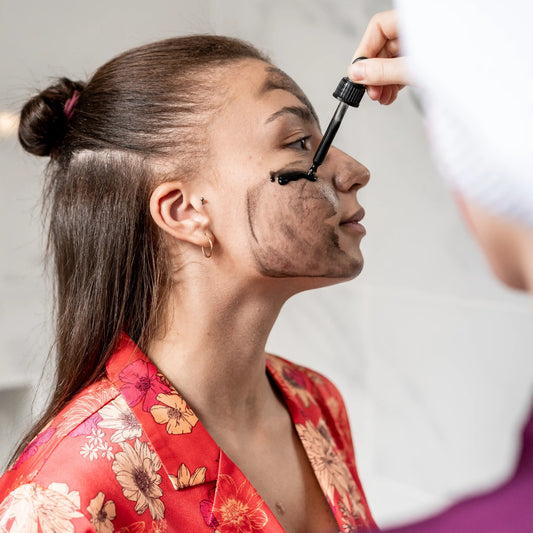 Close-up profile of two women: one applying Clémence & Vivien black peeling mask to the other's cheek and nose with a dropper, the woman receiving the mask wears a red floral robe with her hair pulled back in a bun.