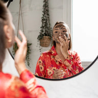Woman applying Clémence et Vivien dark peeling mask to her face while looking in a round bathroom mirror, wearing a red floral robe, with hanging plants and marble tiles in the background.