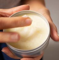 A close-up of a hand scooping Clémence et Vivien Mini Baume Onctueux — Lait d'Amande from an open round metal tin, with fingers blurred in motion.
