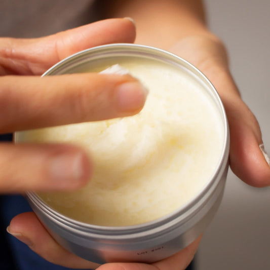 A close-up of a hand scooping a Clémence et Vivien Mini Baume Onctueux Fleurs d'Été from an open round metal tin, with fingers blurred in motion.