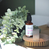 An amber bottle with Clémence et Vivien Sérum perfecteur de peau
sits on a wooden brush with natural bristles, styled beside green eucalyptus leaves on a tabletop.