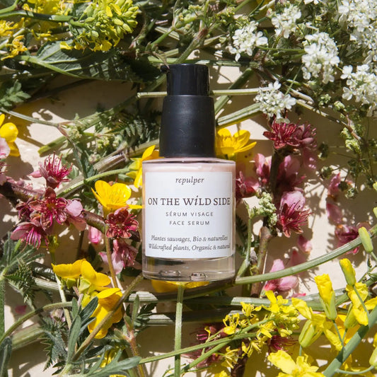 Skincare serum bottle labeled 'On the Wild Side' surrounded by wildflowers on a light background