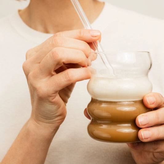 Person holding a glass of layered collagen drink made with Demain Beauty Age with Grace Augmented Collagen, served with a straw against a neutral background.