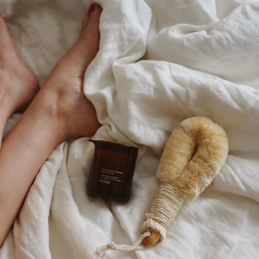 Person lying on a bed next to a massage candle glass jar and natural fiber body brush
