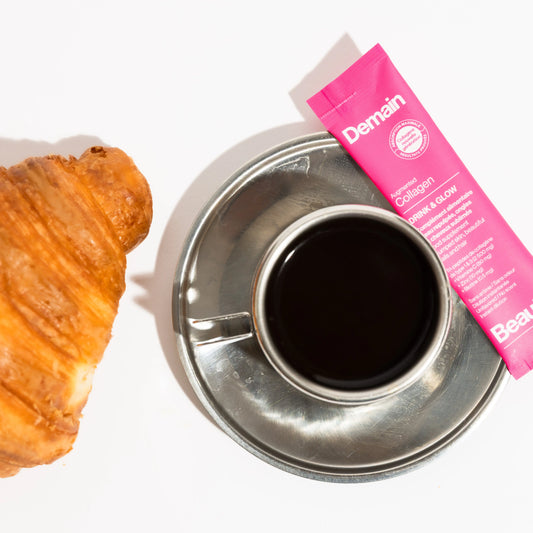 Cup of coffee, croissant and the Drink and Glow collagen sachet from Demain Beauty next to it