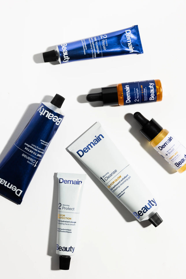 Six Demain Beauty skincare products arranged diagonally on a clean white background, including blue and white tubes and dropper bottles with black caps.