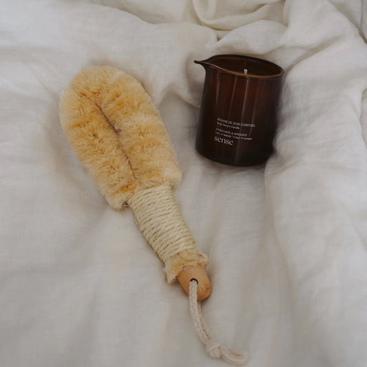 Body brush placed next to an amber glass jar containing a massage candle on a white textured surface.