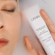 A close-up of a woman’s face surrounded by milky water, holding UMAÏ Hydrating Cream in her hand.