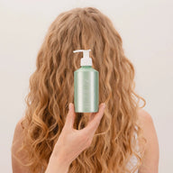 Person holding a green bottle of Lao Care Light Curl Jelly with a pump, positioned beside light curly hair against a plain background