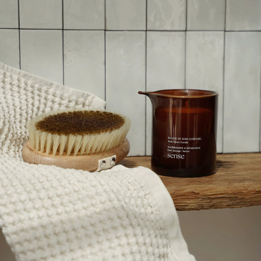 Massage candle in an amber glass jar placed next to a body brush and white towel on a wooden surface, against a tiled wall