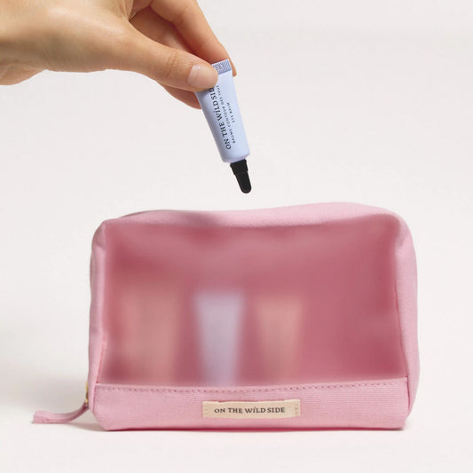 On the Wild Side pink Travel toiletry bag with 4 travel minis inside and a hand holding a mini product over it on a light background