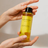 A person's hands holding a clear glass bottle filled with On the Wild Side Huile de Soin Coprs body oil on neutral background 
