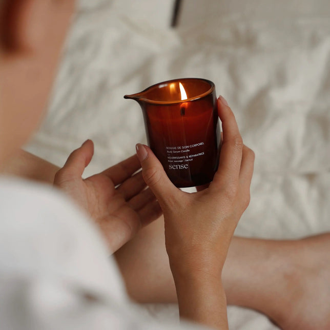 Person holding an amber glass jar containing a nourishing massage candle, with a softly blurred background.