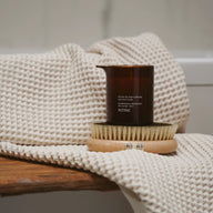 Amber glass jar and body brush placed on a textured towel against a neutral background.