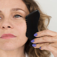 Woman using a black facial gua sha tool on her face against a white background