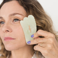 Woman using a green facial jade gua sha on her face against a white background