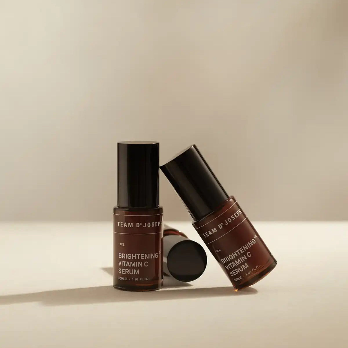 Two bottles of TEAM DR JOSEPH Brightening Vitamin C Serum, one standing and one leaning, with a black cap resting beside them on a beige background
