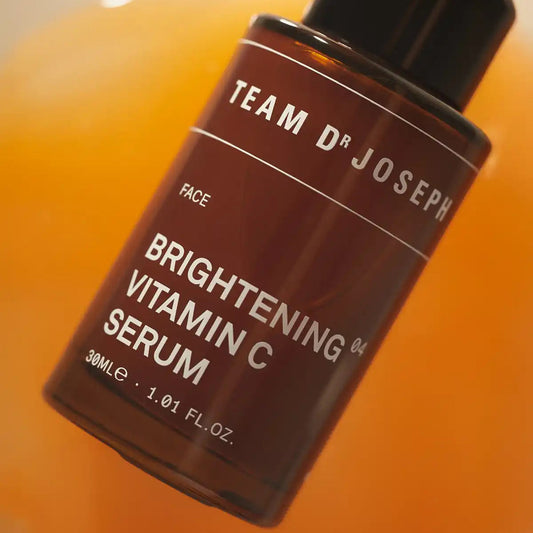 Close-up of TEAM DR JOSEPH Brightening Vitamin C Serum 30 ml amber pump bottle against a pool of orange serum