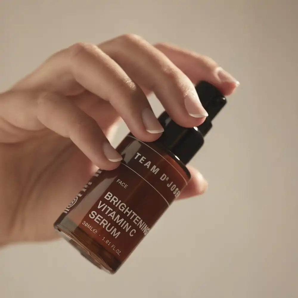Two bottles of TEAM DR JOSEPH Brightening Vitamin C Serum, one standing and one leaning, with a black cap resting beside them on a beige background