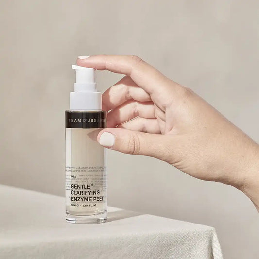 Hand pressing the pump of a TEAM DR JOSEPH Gentle Clarifying Enzyme Peel 50 ml bottle on a linen-covered table