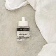 TEAM DR JOSEPH Hyaluronic Repair Booster Serum lying on grey stone beside soft white linen