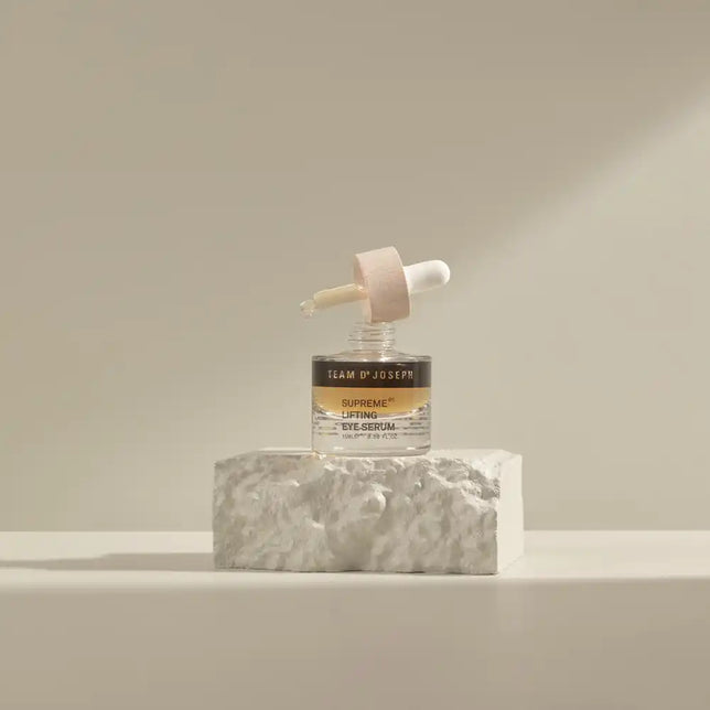 Open bottle of TEAM DR JOSEPH Supreme Lifting Eye Serum with its wooden pipette resting on top, on a white stone block