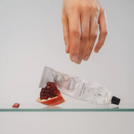 Hand holding a tube of cream above a glass surface with pomegranate seeds.
