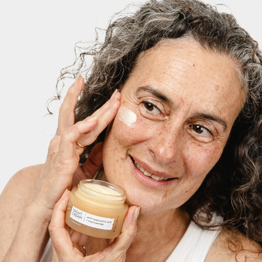 Woman with grey curly hair pressing UpCircle Night Cream onto her cheekbone, holding the 55 ml amber glass jar.